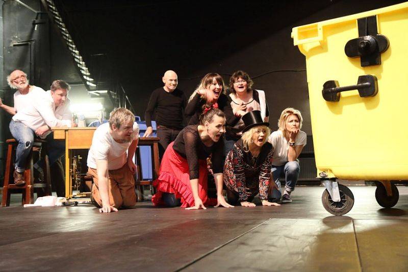 Zdjęcie aktorów podczas spektaklu. Aktorzy z zaciekawieniem i śmiechem stoją i kleczą ,spoglądając w kierunku dużego żółtego kubła na śmieci.