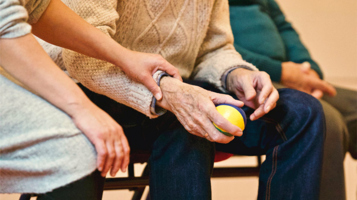 Zdjęcie trzech siedzących starszych osób, kadra na ręcę. Mężczyzna trzyma w ręku małą piłkę.