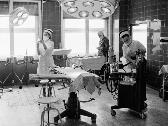Czarno-białe zdjęcie z bloku operacyjnego Szpitala Wojewódzkiego w Tychach z 1973 roku. Na zdjęciu stół, duża lampa oraz trzy pielęgniarki wykunujące różne czynności