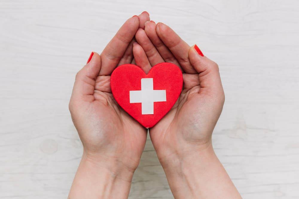Zdjęcie dłoni trzymających czerwone serce ze znakiem krzyża lekarskiego.