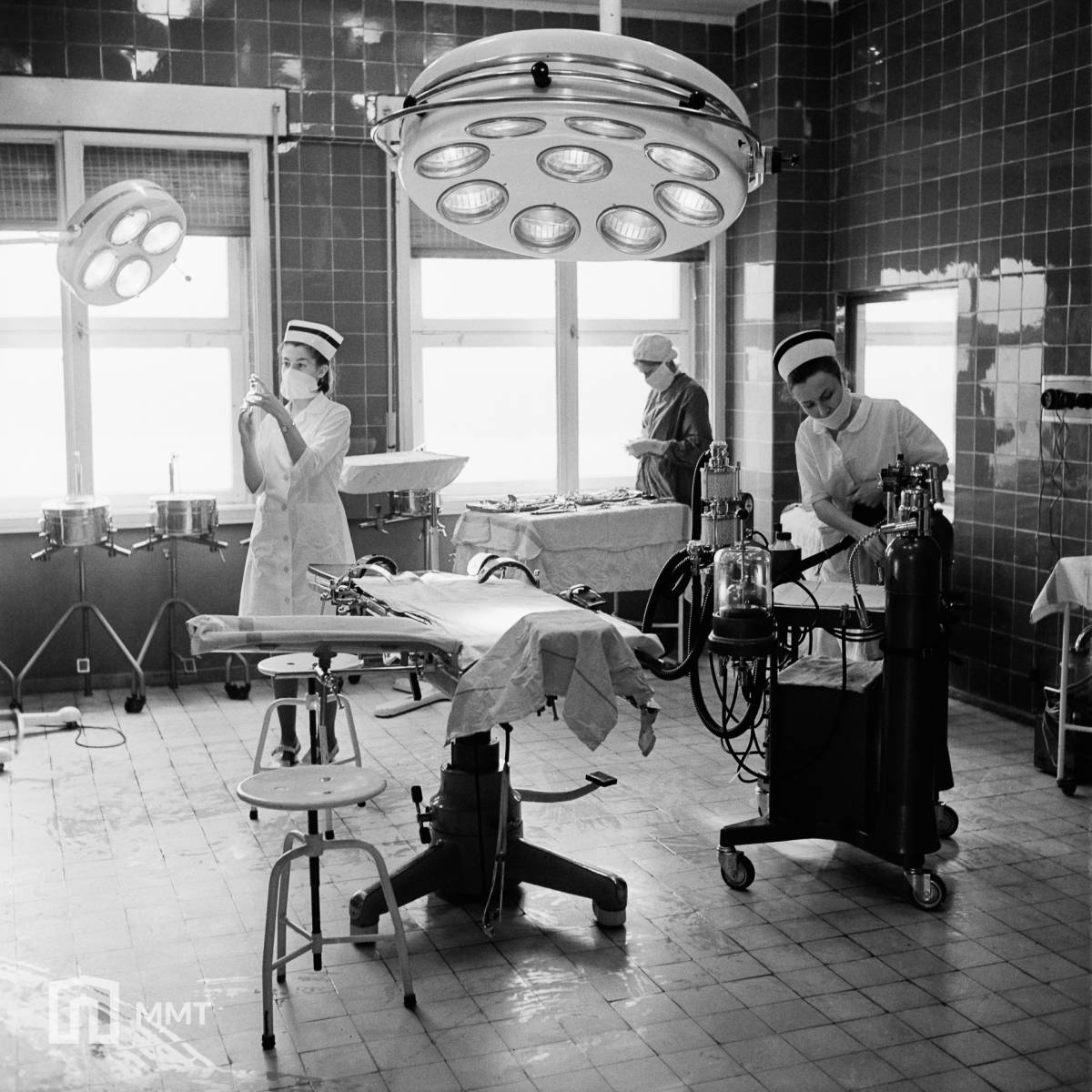 Czarno-białe zdjęcie z bloku operacyjnego Szpitala Wojewódzkiego w Tychach z 1973 roku. Na zdjęciu stół, duża lampa oraz trzy pielęgniarki wykunujące różne czynności
