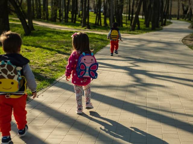 Dzieci w wieku przedszkolnym z kolorowymi plecakami, idące parkową alejką