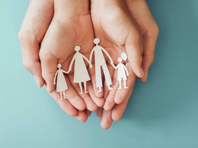 Wewnętrzna strona złączonych dłoni osoby dorosłej, w niej złączone dłonie dziecka - na dłoniach dziecka wycinanka - postacie tworzące rodzinę.