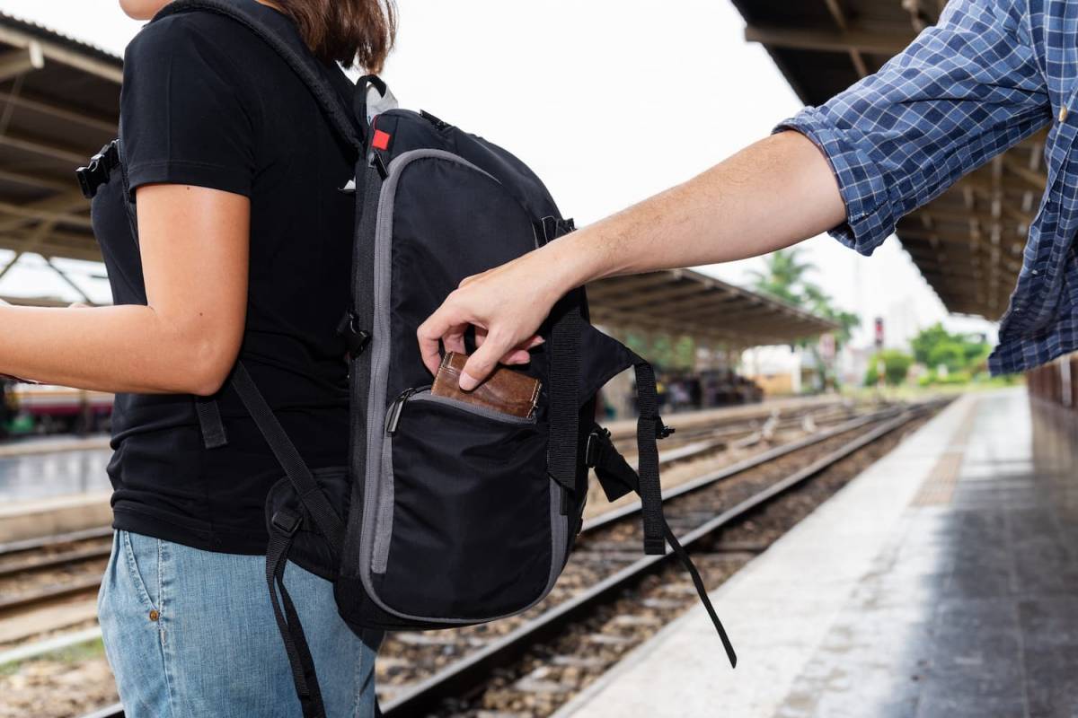 Męska dłoń sięgająca do plecaka kobiety stojącej na peronie kolejowym.