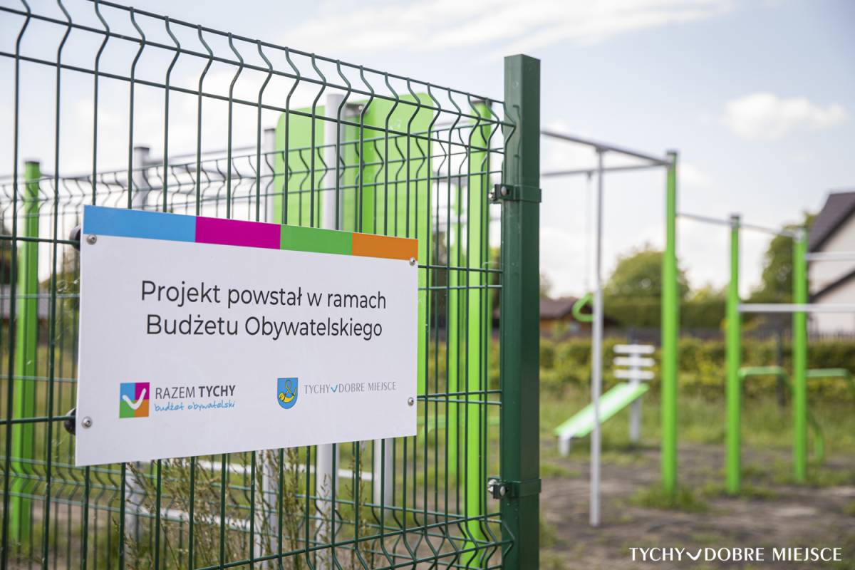 Tabliczka na ogrodzeniu, z tyłu widać plac zabaw zrealizowany w ramach Budżetu Obywatelskiego