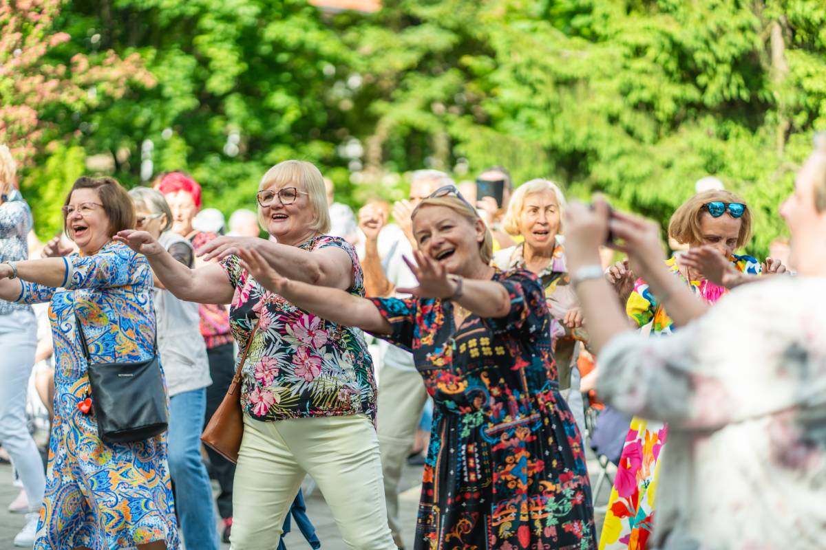 Trzy uśmiechnięte seniorki tańczą z wyciągniętymi rękami, w tle widać tłum ludzi.