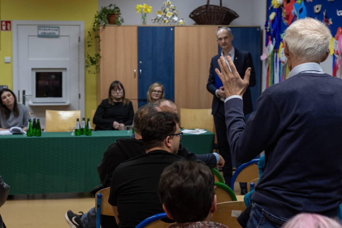 Trzy kobiety siedzą, jeden mężczyzna stoi przodem do mieszkańców. Tyłem do zdjęcia siedzi grupa mieszkańców, jeden starszy mężczyzna stoi z podniesioną ręką.