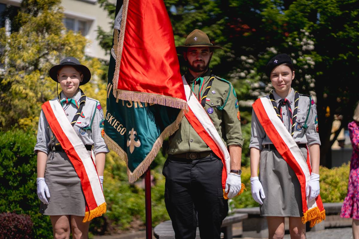 Mężczyzna ze sztandarem oraz dwie kobiety w biało-czerwonych szarfach stoją na baczność na tle krzewów i budynku.