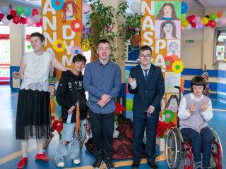 Czworo dzieci i jedna dziewczynka na wózku uśmiechają się do zdjęcia na korytarzu ozodbionym balonami i rysunkami.