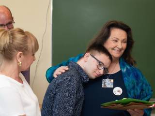 Chłopiec przytula się do uśmiechniętej kobiety, obok nich stoi mężczyzna i kobieta, w tle tablica szkolna. 