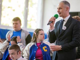 Mężczyzna mówi do mikrofonu, wokół niego stoją dzieci, w tle okna sali gimnastycznej.