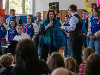 Kobieta z mikrofonem przemawia do grupy dzieci na sali gimnastycznej.