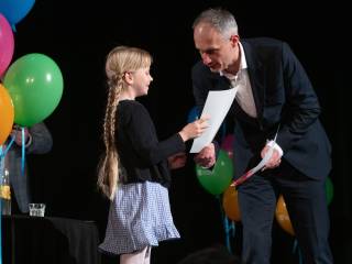 Mężczyzna na scenie wręcza nagrodę dziewczynce, w tle widać balony.