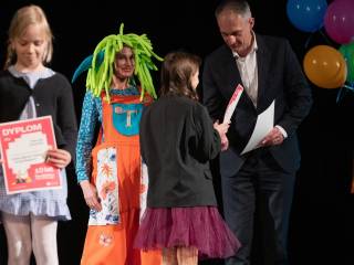 Cztery osoby stoją na scenie. Mężczyzna wręcza nagrodę dziewczynce, obok nich stoi przebrana aktorka, z lewej strony dziewczynka wraca z nagrodą. 