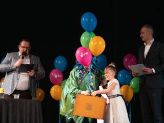 Dwóch mężczyzn stoi na scenie, jeden mówi do mikrofonu, obok nich stoi kobieta i dziewczynka, która odbiera nagrodę.