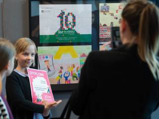 Kobieta robi zdjęcie dziewczynce z dyplomem na tle prac konkursowych.