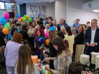 Duża grupa ludzi zgromadzona wokół stołu z tortem, pomiedzy nimi znajdują się balony. 