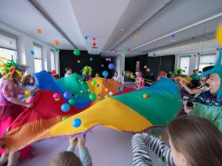 Grupa dzieci, rodzice i przebrane aktorki podrzucają kolorową chustą animacyjną piłeczki w sali teatralnej.