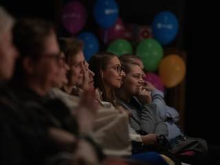 Grupa widzów ogląda przedstawienie teratralne, w tle widać balony.