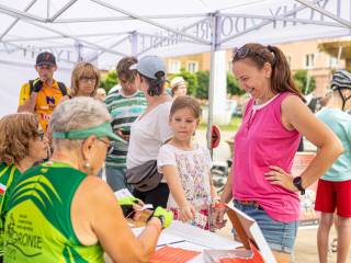 Pod białym namiotem plenerowym siedzi starsza kobieta w zielonej sportowej bluzce przodem do stolika; po drugiej stronie stoją kobieta w różowej koszulce oraz dziewczynka. W tle widać osoby czekające w kolejce i fragment miejskiej zabudowy.