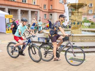 Mężczyzna i chłopiec jadą na rowerach. W tle widać fontannę, namiot plenerowy, baner z napisem "Metrogra", ludzi i budynki.