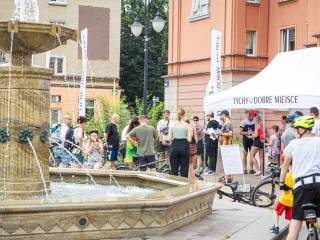 Fontanna, obok której stoją ludzie z rowerami. W tle widać dwa budynki oraz biały namiot plenerowy z napisem "Tychy dobre miejsce".