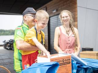 Mężczyzna trzyma na rękach chłopczyka, który sięga ręką do kartonu, obok stoi uśmiechnięta kobieta w sukience. W tle widać budynek i jezioro.