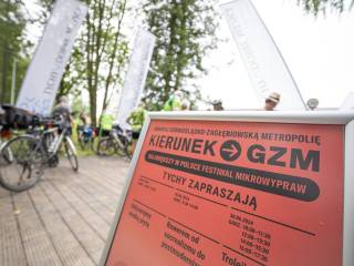 Stojak reklamowy z pomarańczową kartką z napisem "Kierunek GZM-Tychy zapraszają". W tle widać ludzi z rowerami, drzewa i białe banery. 