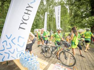 Grupa osób z rowerami stoi na utwardzonym podłożu z drewnianych paneli, otoczonym trawą, na pierwszym planie widać stolik z butelkami, obok niego stoi baner z napisem "Tychy". W tle znajduje się las.
