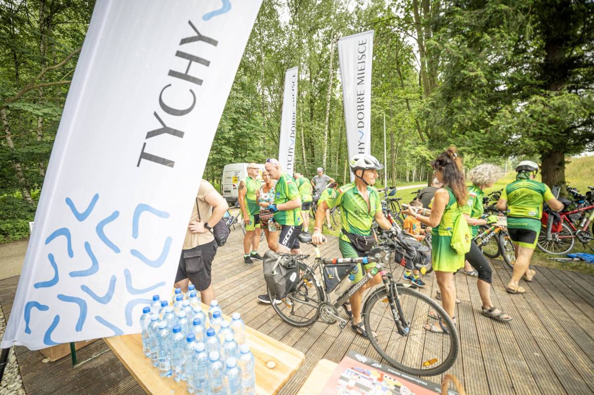 Grupa osób z rowerami stoi na utwardzonym podłożu z drewnianych paneli, otoczonym trawą, na pierwszym planie widać stolik z butelkami, obok niego stoi baner z napisem "Tychy". W tle znajduje się las.