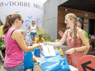 „Kobieta w różowym ubraniu podaje kartkę stojącej naprzeciw kobiecie i dziecku. Obok nich znajdują się ludzie, w tle widać trawę i budynek.