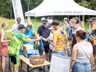 Starszy mężczyzna w zielonym sportowym stroju i kobieta w różowym ubraniu stoją przed stolikiem i rozdają ludziom niebieskie torby. W tle biały namiot plenerowy i park.
