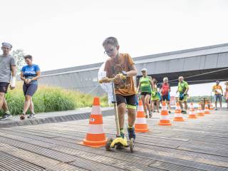 Chłopiec jedzie na hulajnodze po drewnianym podłożu, mijając pomarańczowe pachołki; w tle widać ludzi, trawę, budynek i jezioro.
