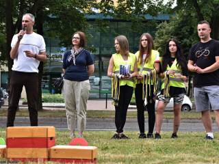 Na tle ulicy i nowoczesnego przeszklonego budynku stoi sześć osób. Mężczyzna mówi do mikrofonu, obok niego kobieta, trzy dziewczyny z medalami i drugi mężczyzna.