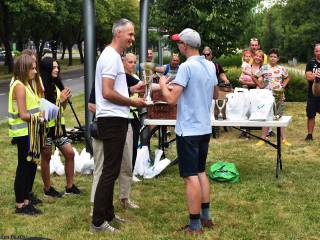 Mężczyzna wręcza puchar innemu mężczyźnie, w tle widoczni są ludzie, stolik z nagrodami i park