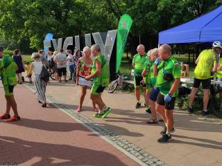 Duża grupa ludzi w sportowych strojach stoi na deptaku przy namiocie i napisie "Tychy", w tle widać park.