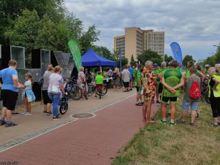 Duża grupa ludzi w sportowych strojach stoi na ścieżce przy namiocie i napisie "Tychy", w tle widać park i budynek Urzędu Miasta.