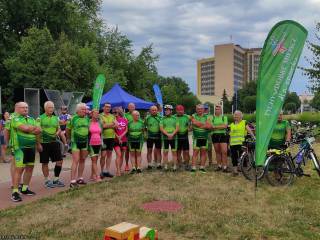 Duża grupa ludzi w sportowych strojach stoi na trawie przy drodze dla pieszych i uśmiecha się do zdjęcia, w tle fragment napisu "Tychy" i budynek Urzędu Miasta.