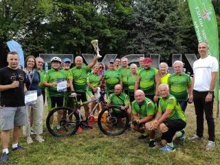 Duża grupa ludzi w sportowych strojach uśmiecha się do zdjęcia, w tle napis "Tychy" i zielone drzewa.