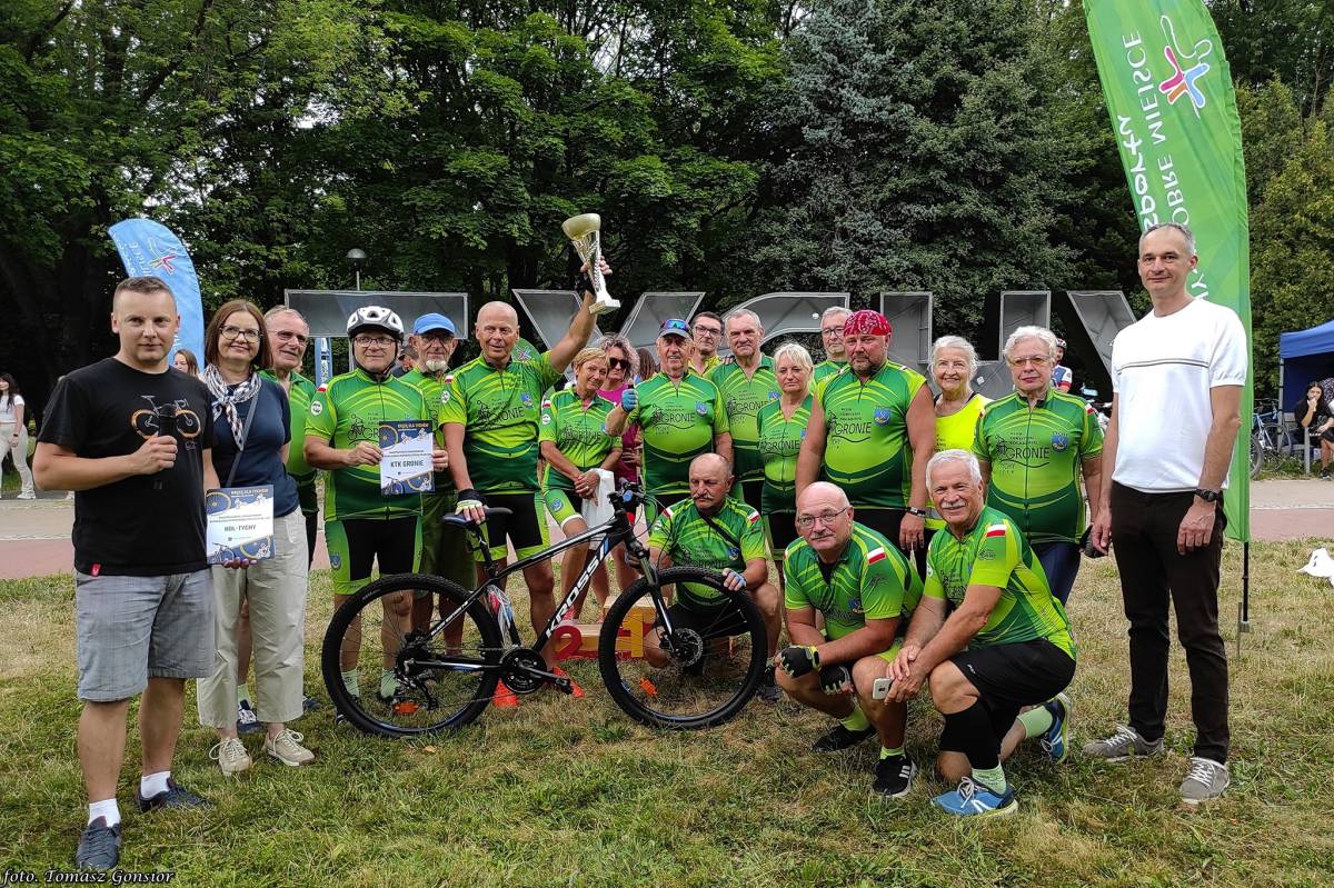 Duża grupa ludzi w sportowych strojach uśmiecha się do zdjęcia, w tle napis "Tychy" i zielone drzewa.