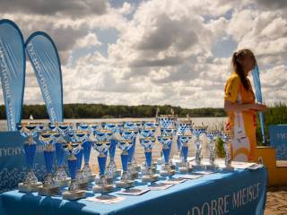 Kobieta w żółtym stroju sportowym stoi przy stoliku z napisem "Tychy dobre miejsce", na którym leżą niebieskie puchary, w tle jezioro.