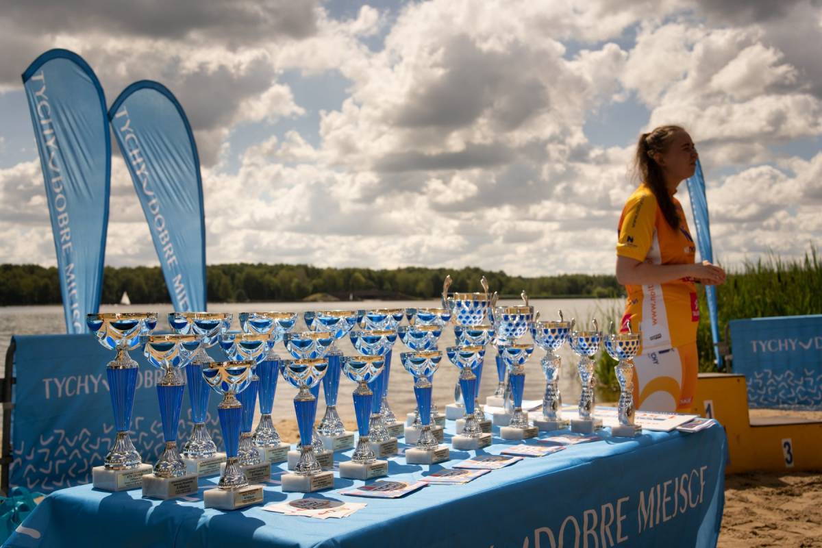 Kobieta w żółtym stroju sportowym stoi przy stoliku z napisem "Tychy dobre miejsce", na którym leżą niebieskie puchary, w tle jezioro.