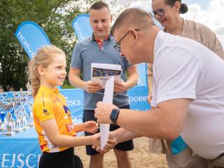 Mężczyzna w białej koszulce wręcza dyplom dziewczynce w sportowym żółtym stroju, w tle widać mężczyznę, kobietę i puchary, które leżą na stoliku.