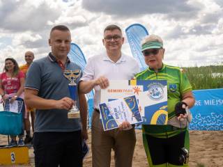 Trzy osoby stoją i uśmiechają się do zdjęcia. Mężczyzna po lewej trzyma puchar, mężczyzna w środku oraz kobieta w stroju sportowym wspólnie trzymają dyplom i nagrodę. W tle widoczni są ludzie i jezioro.