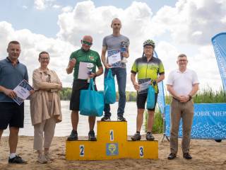 Sześć osób stoi i uśmiecha się do zdjęcia. Na środku, na podium, trzech mężczyzn trzyma nagrody. Po lewej stronie stoi mężczyzna z dyplomem oraz kobieta, a po prawej — kolejny mężczyzna. W tle widoczne jest jezioro.