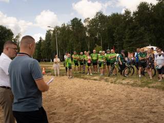 Dwaj mężczyźni stoją plecami do aparatu i rozmawiają. Przed nimi rozciąga się pas piasku, a dalej na trawie stoi grupa osób.