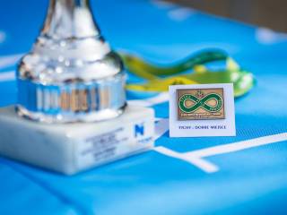 Na niebieskim tle znajduje się srebrny puchar oraz odznaka z napisem "Tyska Pętla Rowerowa".