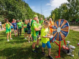 Grupa dorosłych i dzieci stoi w kolejce do zakręcenia kołem, w tle las.