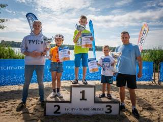 Troje dzieci — dziewczynka i dwóch chłopców — stoi na podium z nagrodami. Po obu stronach znajdują się dwaj mężczyźni. W tle widoczne jest jezioro.