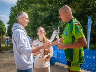 Kolarz w sportowym stroju odbiera dyplom i puchar od mężczyzny i kobiety, w tle znajdują się ludzie i las.